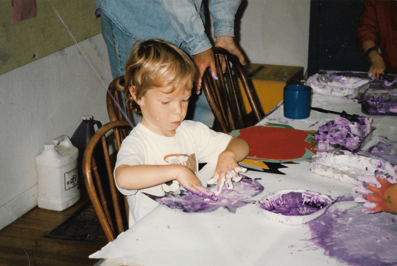 A young blond-haired boy mixing purple paint with his hands at a messy table covered in arts and craft supplies.