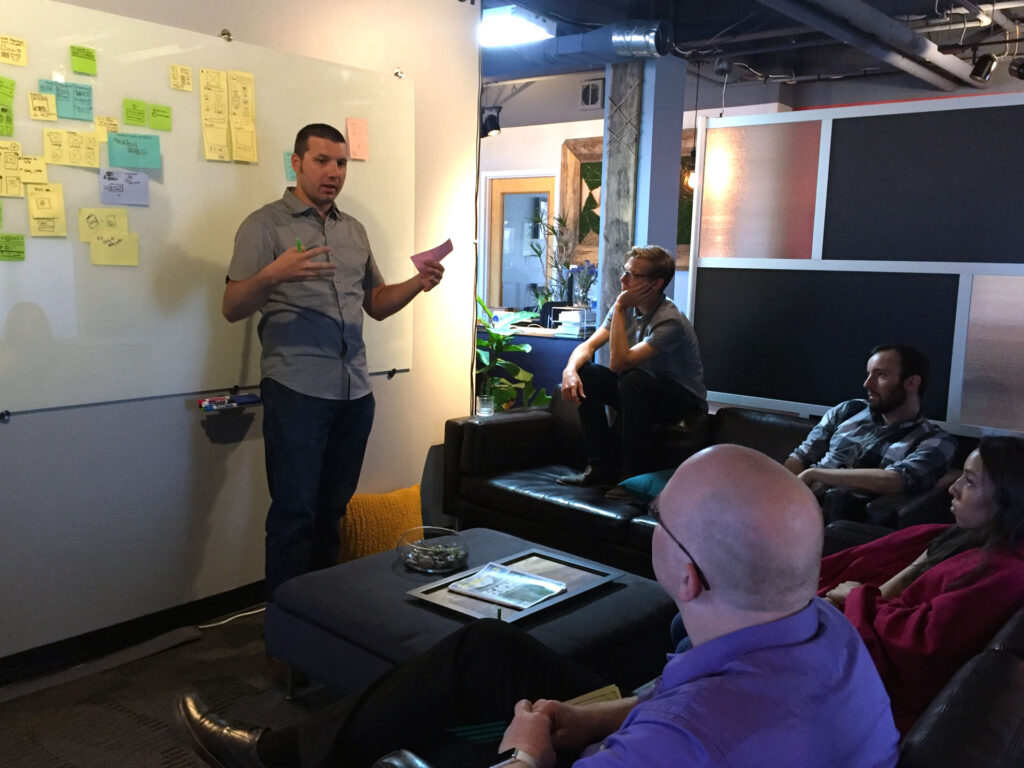 ImageQuix’s Craig Valente presents some of his ideas to the project team. He stands in front of a whiteboard adorned with sticky notes, his teammates seated on comfy couches.