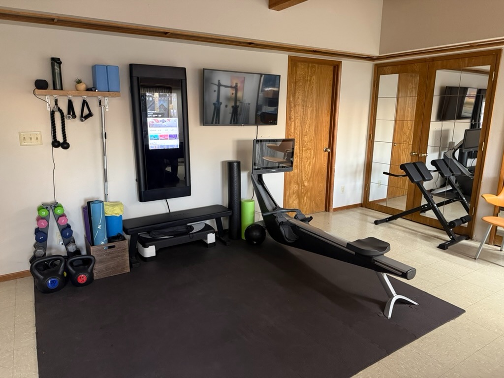 A photo of our home gym. On the wall is a Tonal system for weight lifting. It is stored away at the moment and looks like a vertical TV. Hanging to the left of the Tonal from the bottom of a shelf are its accessories: a rope, two handles, and bar. To the right of the Tonal is an actual TV. Below it is a Hydrow rowing machine. Around the room are various other workout accessories like dumbbells. The closet doors have mirrors on them.
