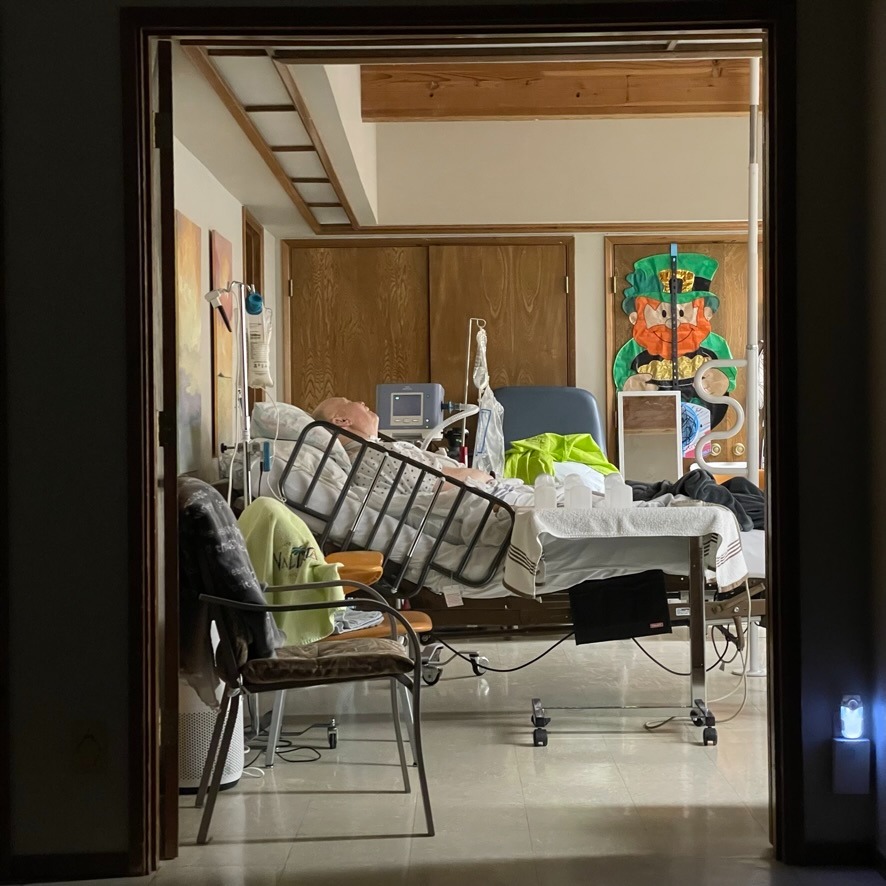My father is asleep in a hospital bed in our house. In the foreground, a bag of food hangs off a pole where a machine slowly provided food via a  gastrostomy tube. In the background, a ventilator acts as a BiPap connected to his trach at night. A large leprechaun decoration hangs on the wall. My dad once joked the leprechaun was his childhood friend Gary and after that Gary stayed on the wall year round.